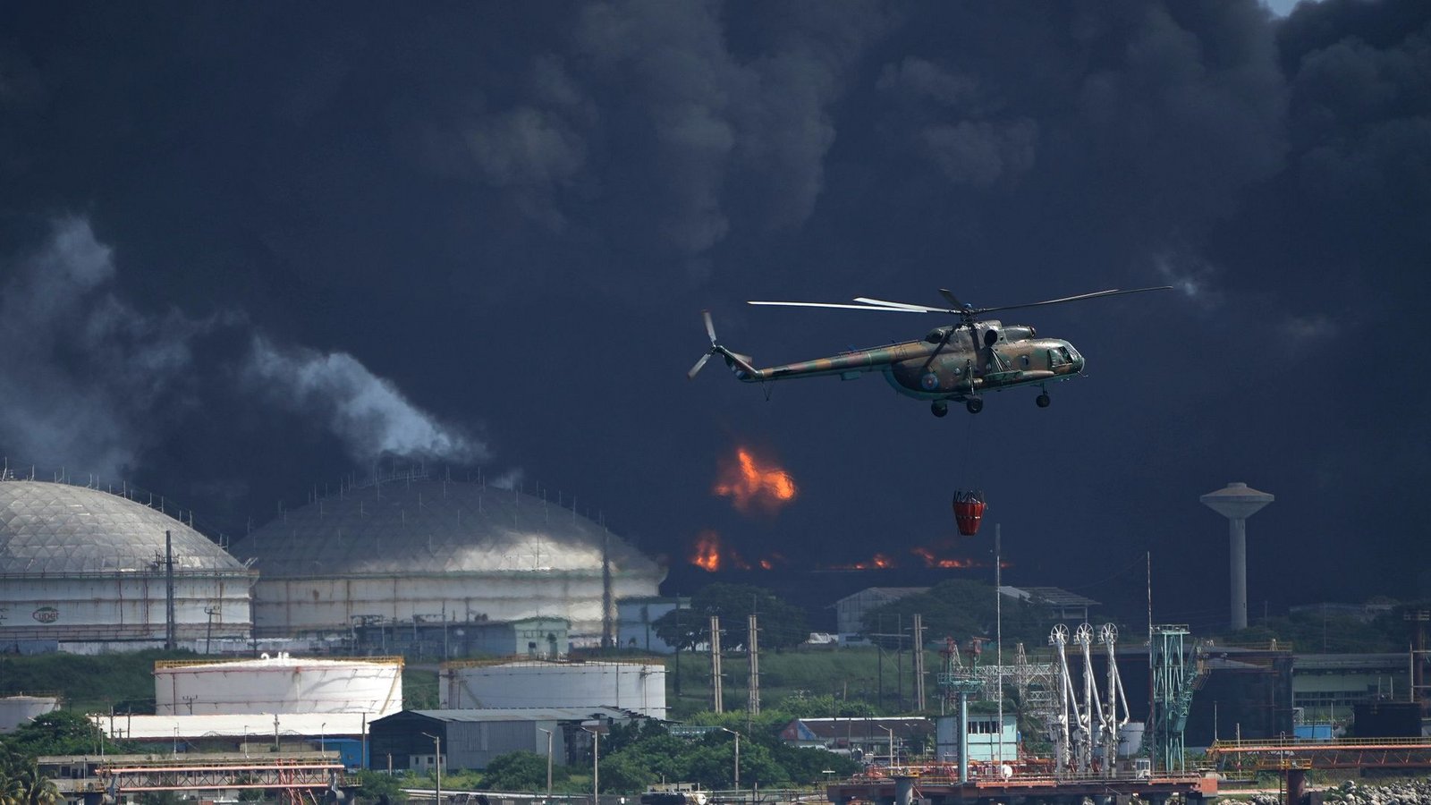 Ein mit Wasser beladener Hubschrauber fliegt über die Supertanker-Basis Matanzas, von der dunkler Rauch und Flammen aufsteigen, während Feuerwehrleute versuchen, den Brand zu löschen.Foto: Ramon Espinosa/AP/dpa