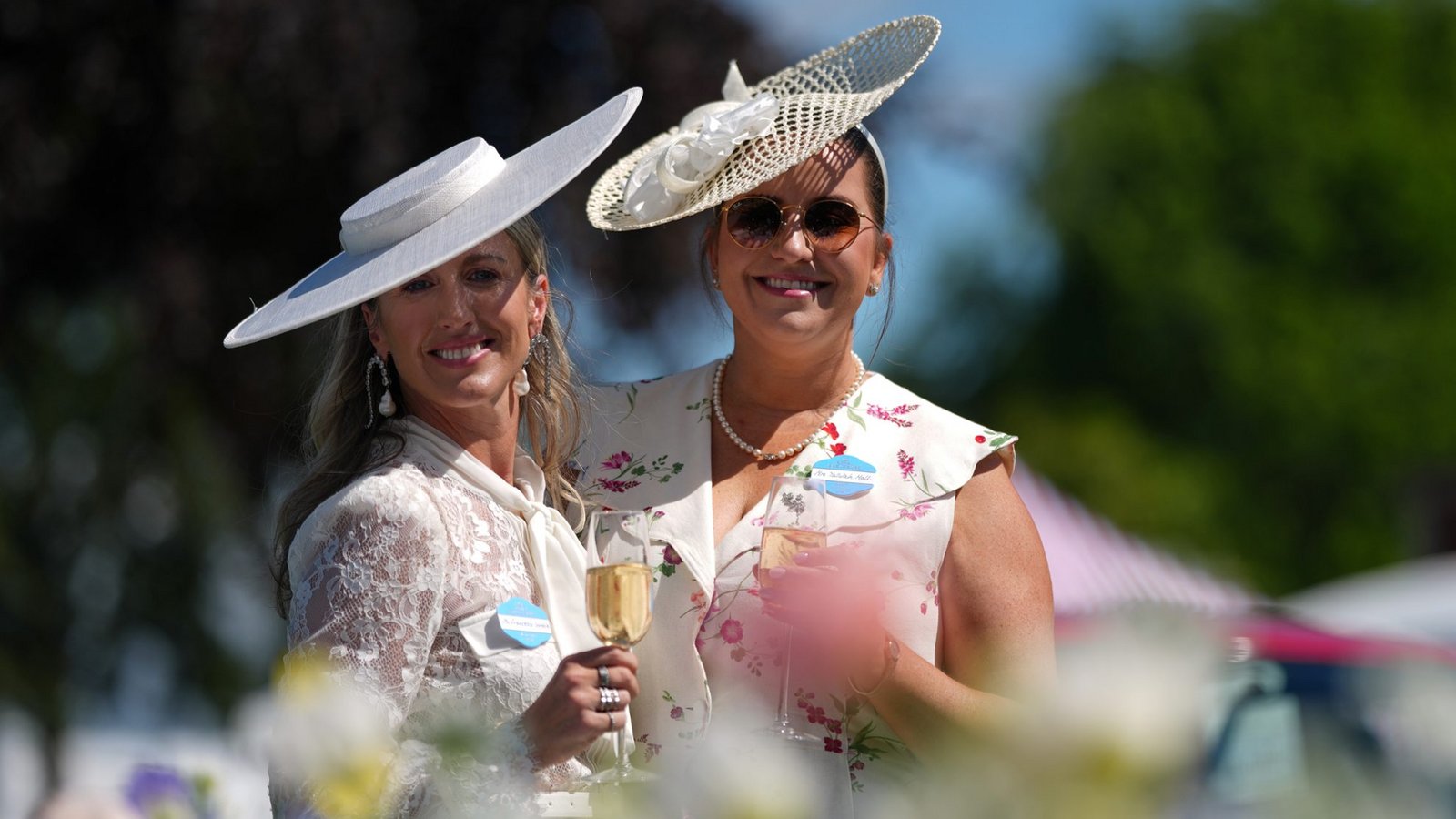 Yallulah Hall und Francesca Gamble (l) zeigen zurückhaltendere Sommerlooks in hellen Tönen: ein weißer Tellerhut zur Spitzenbluse und ein cremefarbener Schräghut mit Blütendetail zum floralen Kleid. Perlen und Champagnerglas unterstreichen den klassischen Ascot-Stil.Foto: picture alliance/dpa/AP | Alastair Grant