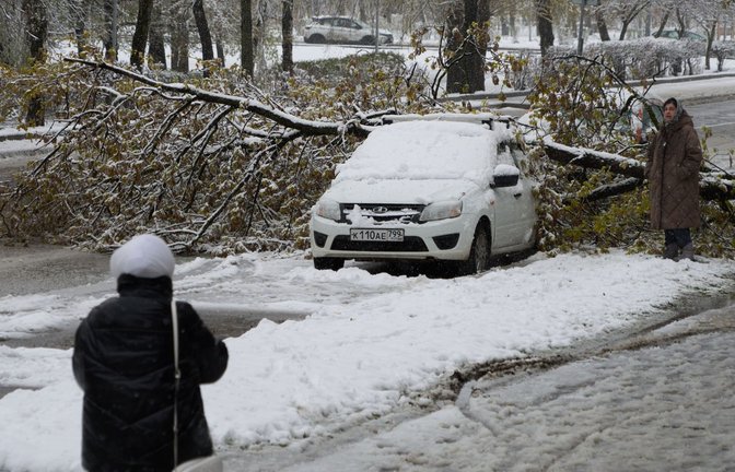 Eine Frau betrachtet ein Auto, das nach einem Schneefall unter einem Baum eingeklemmt ist.<span class='image-autor'>Foto: Alexander Zemlianichenko/AP/dpa/Alexander Zemlianichenko</span>