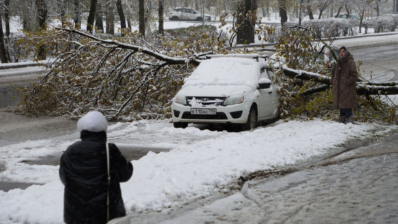 Eine Frau betrachtet ein Auto, das nach einem Schneefall unter einem Baum eingeklemmt ist.Foto: Alexander Zemlianichenko/AP/dpa/Alexander Zemlianichenko Eine Frau betrachtet ein Auto, das nach einem Schneefall unter einem Baum eingeklemmt ist.Foto: Alexander Zemlianichenko/AP/dpa/Alexander Zemlianichenko