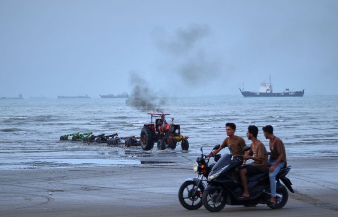 Junge Männer fahren mit Motorrädern am Ufer entlang, während ein Traktor Geräte aus dem Wasser zieht und Handelsschiffe in der Straße von Hormuz vor Anker liegen. (zu dpa: «Rund 400 Schiffe stecken noch im Persischen Golf fest»)<span class='image-autor'>Razieh Poudat/ISNA/AP/dpa</span>