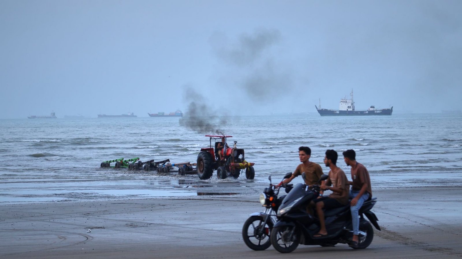 Junge Männer fahren mit Motorrädern am Ufer entlang, während ein Traktor Geräte aus dem Wasser zieht und Handelsschiffe in der Straße von Hormuz vor Anker liegen. (zu dpa: «Rund 400 Schiffe stecken noch im Persischen Golf fest»)<span class='image-autor'>Razieh Poudat/ISNA/AP/dpa</span>