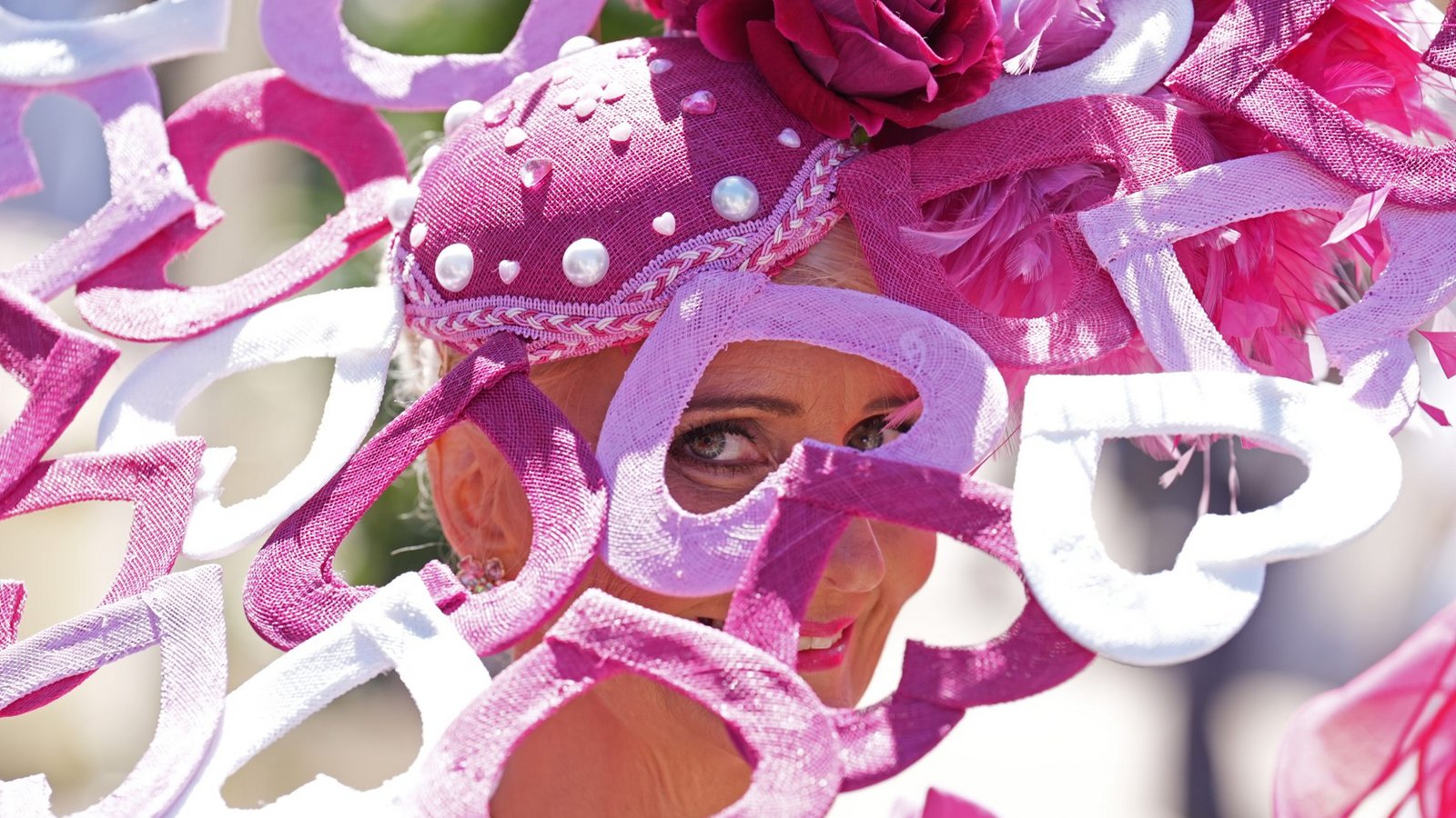 Eine Besucherin von Royal Ascot trägt einen rosafarbenen Hut mit Perlen, Blüte und Federn. Besonders auffällig sind die ausladenden, geschwungenen Elemente in Pink, Rosa und Weiß, die den Kopfschmuck fast wie eine textile Skulptur wirken lassen.Foto: picture alliance/dpa/PA Wire | Mike Egerton