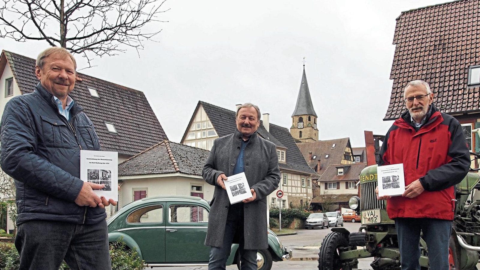 Heinz Burkhardt (links) und Edgar Winkler (rechts) übergeben Ortsvorsteher Rolf Allmendinger ihr Buch. Mit dabei zwei „Zeitzeugen“: ein VW Käfer und ein Deutz-Traktor von 1959.