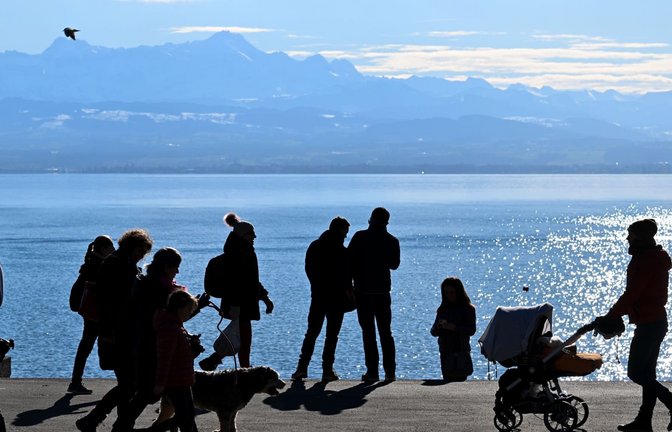 Mehrere Menschen laufen an der Uferpromenade entlang, während im Hintergrund der Bodensee und die Alpen in der Schweiz zu sehen sind. (zu dpa: «Hochalpiner Ausflug mit Kinderwägen: 36 Personen gerettet»)<span class='image-autor'>Felix Kästle/dpa</span>