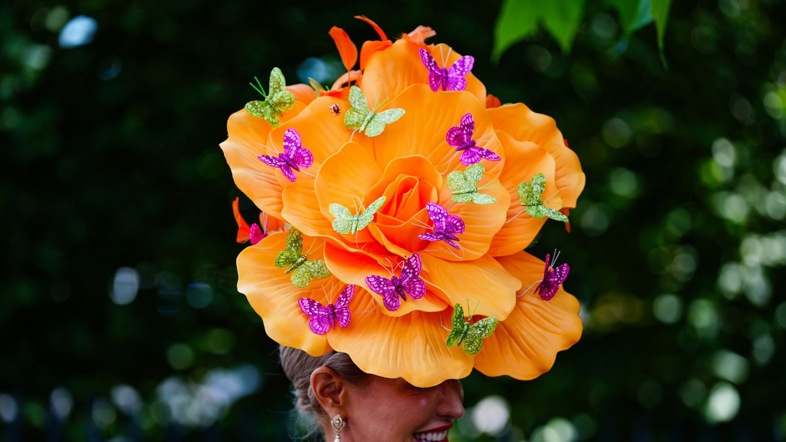 Eine Besucherin von Royal Ascot trägt einen übergroßen orangefarbenen Blütenhut, der mit violetten und grünen Schmetterlingen verziert ist. Der florale Kopfschmuck wird mit Perlenkette, Tropfenohrringen und einem farbenfrohen Kleid kombiniert.Foto: picture alliance/dpa/PA Wire | David Davies