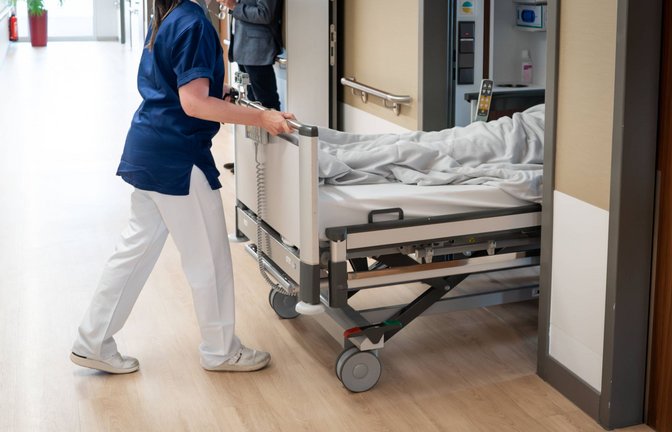 Eine Pflegekraft schiebt einen Patienten im Krankenbett in ein Krankenzimmer (Symbolbild).<span class='image-autor'>Foto: IMAGO/Political-Moments/IMAGO</span>