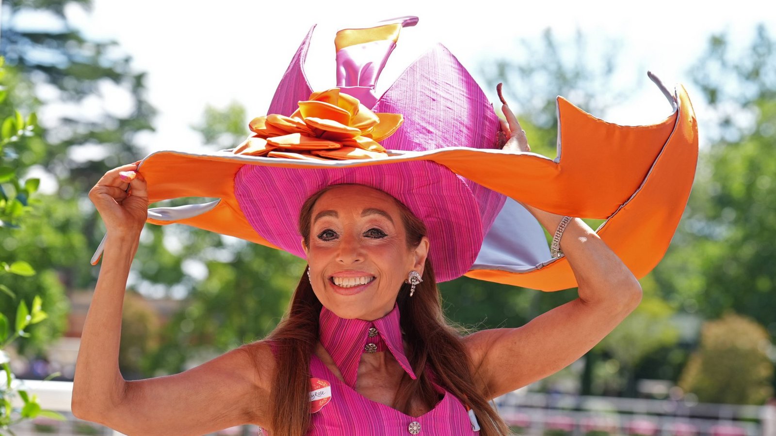 Eine Besucherin von Royal Ascot trägt einen extravaganten Hut in Pink und Orange mit weit ausladender, geschwungener Krempe und Blütendetail. Das knallige Pink des Kopfschmucks wird im ärmellosen Kleid mit hohem Kragen wieder aufgegriffen.Foto: picture alliance/dpa/PA Wire | Mike Egerton