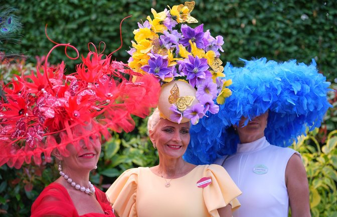 Drei Besucherinnen von Royal Ascot setzen auf maximal auffälligen Kopfschmuck: ein roter Fascinator mit Federn und floralen Elementen, ein hoher Blumenhut in Gelb und Violett sowie ein voluminöser blauer Federhut zum schlichten weißen Kleid.<span class='image-autor'>Foto: picture alliance/dpa/AP | Kin Cheung</span>