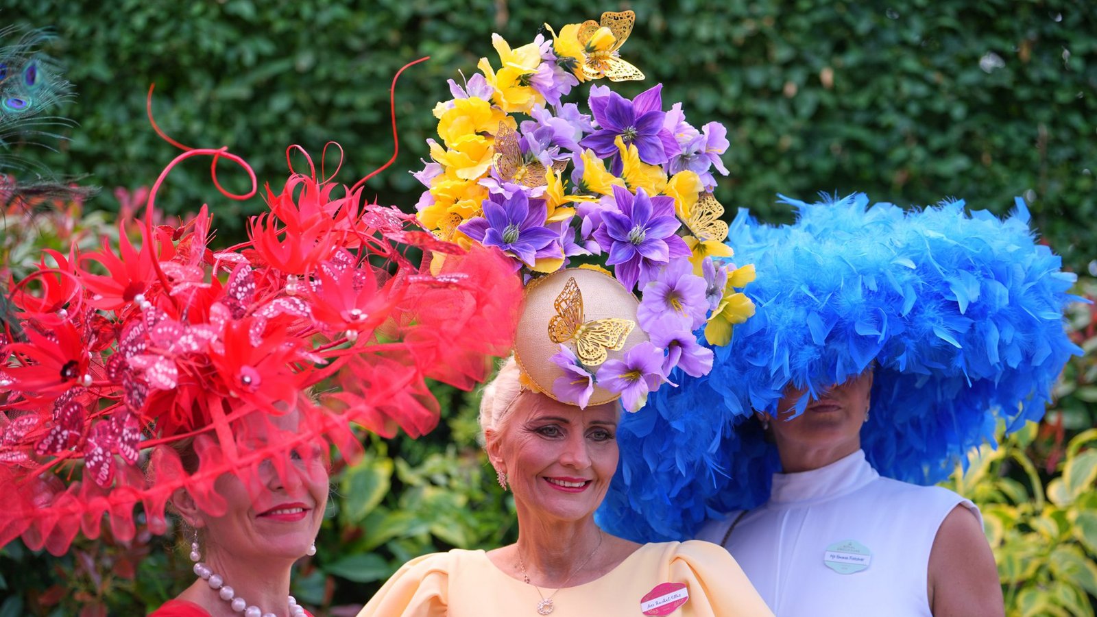 Drei Besucherinnen von Royal Ascot setzen auf maximal auffälligen Kopfschmuck: ein roter Fascinator mit Federn und floralen Elementen, ein hoher Blumenhut in Gelb und Violett sowie ein voluminöser blauer Federhut zum schlichten weißen Kleid.Foto: picture alliance/dpa/AP | Kin Cheung