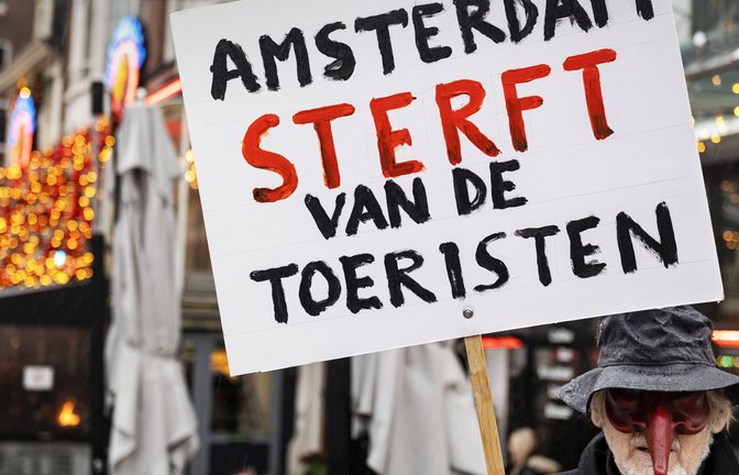 Ein niederländischer Aktivist mit Narrenmaske hält ein  Schild hoch mit der übersetzten Aufschrift: „Amsterdam stirbt an den Touristen“<span class='image-autor'>Foto: imago/Richard Wareham</span>