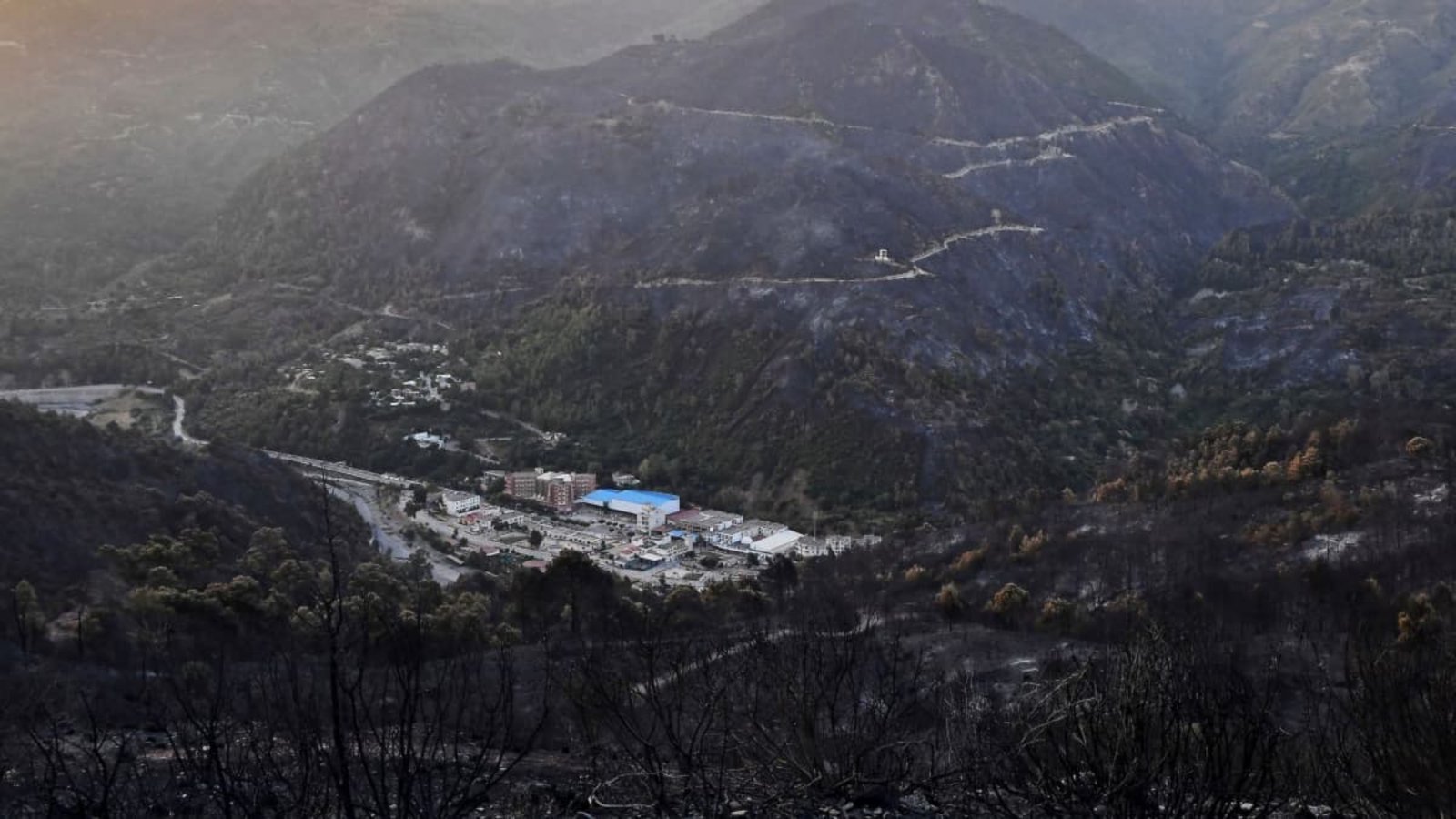 Nach einem Waldbrand umgeben verkohlte Berge ein Dorf im Tal.Fateh Guidoum/AP/dpa Nach einem Waldbrand umgeben verkohlte Berge ein Dorf im Tal.Fateh Guidoum/AP/dpa