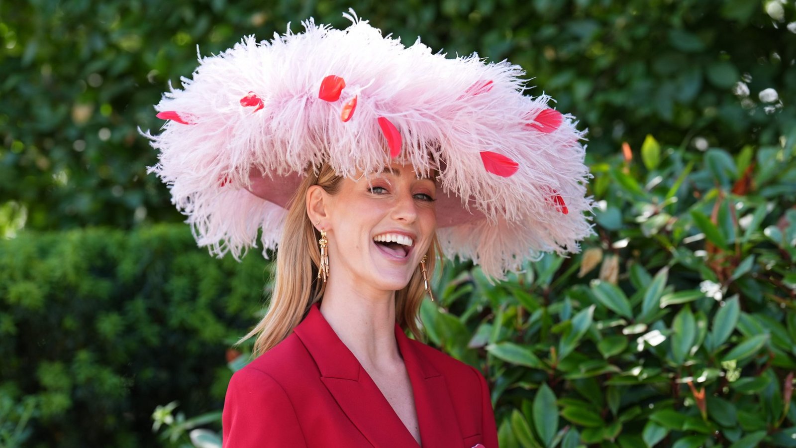 Eine Besucherin von Royal Ascot kombiniert einen roten Hosenanzug mit einem großen, hellrosa Federhut. Rote Blütenblätter setzen farbliche Akzente und greifen den Ton des Outfits wieder auf.Foto: picture alliance/dpa/PA Wire | Mike Egerton