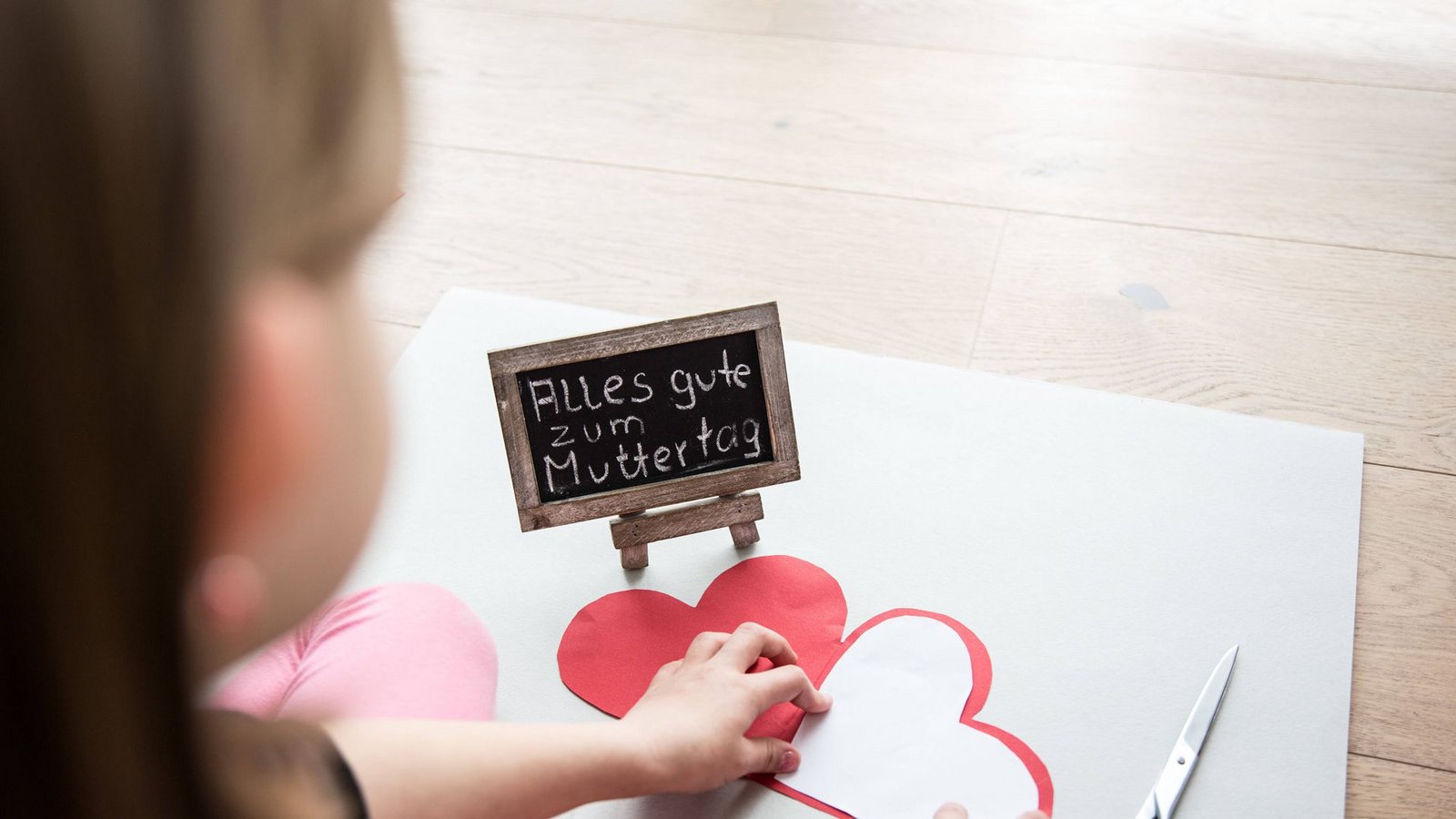 Ein kleines Mädchen sitzt auf einem Laminatfussboden und bastelt zum Muttertag für die Mutter ein Herz aus Pappe als Geschenk, daneben liegt eine Schere. Auf einer Tafel steht mit Kreide geschrieben, alles gute zum Muttertag.Foto: Imago/Fotostand Ein kleines Mädchen sitzt auf einem Laminatfussboden und bastelt zum Muttertag für die Mutter ein Herz aus Pappe als Geschenk, daneben liegt eine Schere. Auf einer Tafel steht mit Kreide geschrieben, alles gute zum Muttertag.Foto: Imago/Fotostand