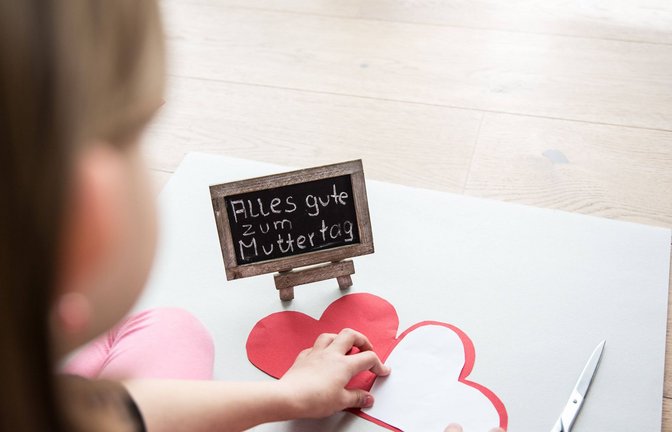 Ein kleines Mädchen sitzt auf einem Laminatfussboden und bastelt zum Muttertag für die Mutter ein Herz aus Pappe als Geschenk, daneben liegt eine Schere. Auf einer Tafel steht mit Kreide geschrieben, alles gute zum Muttertag.<span class='image-autor'>Foto: Imago/Fotostand</span>