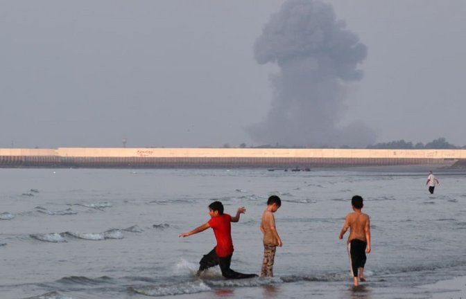 Drei Jungen spielen im seichten Wasser der Straße von Hormus, während im Hintergrund vor der Küste von Bandar Abbas im Iran eine Rauchwolke nach einer Explosion aufsteigt.<span class='image-autor'>Razieh Poudat/ISNA via AP/dpa</span>