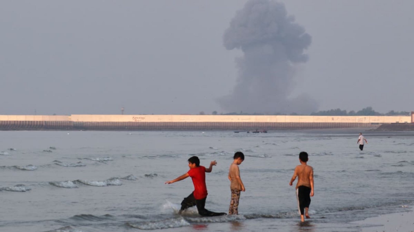 Drei Jungen spielen im seichten Wasser der Straße von Hormus, während im Hintergrund vor der Küste von Bandar Abbas im Iran eine Rauchwolke nach einer Explosion aufsteigt.Razieh Poudat/ISNA via AP/dpa Drei Jungen spielen im seichten Wasser der Straße von Hormus, während im Hintergrund vor der Küste von Bandar Abbas im Iran eine Rauchwolke nach einer Explosion aufsteigt.Razieh Poudat/ISNA via AP/dpa