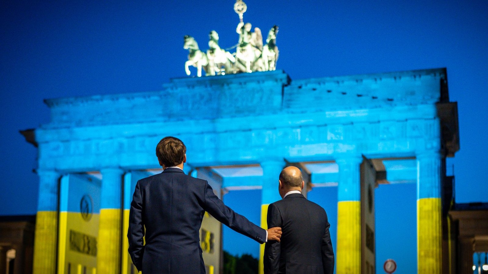 Bundeskanzler Olaf Scholz (r) und Frankreichs Präsident Emmanuel Macron blicken auf das Brandenburger Tor, das in den ukrainischen Nationalfarben erstrahlt.Foto: Michael Kappeler/dpa