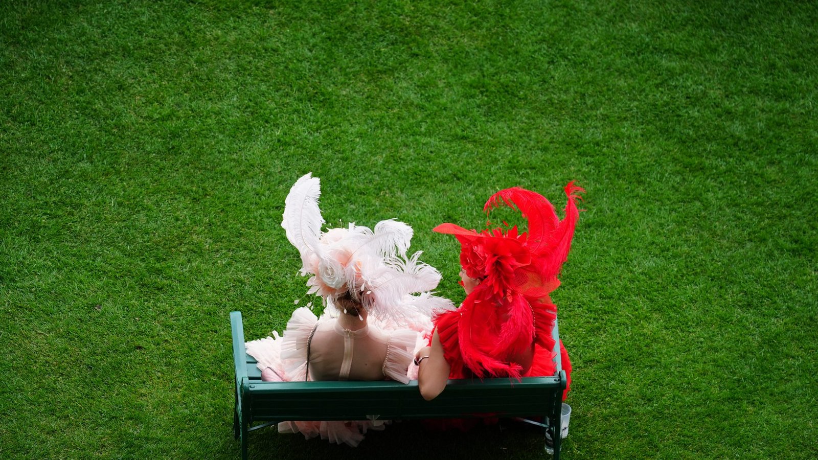 Zwei Besucherinnen von Royal Ascot sitzen in farblich abgestimmten Feder-Looks auf einer Bank: links ein zarter roséfarbener Kopfschmuck mit großen Federn und Tüll, rechts ein dramatischer roter Hut mit ausladenden Federn und passendem Kleid.Foto: picture alliance/dpa/PA Wire | David Davies